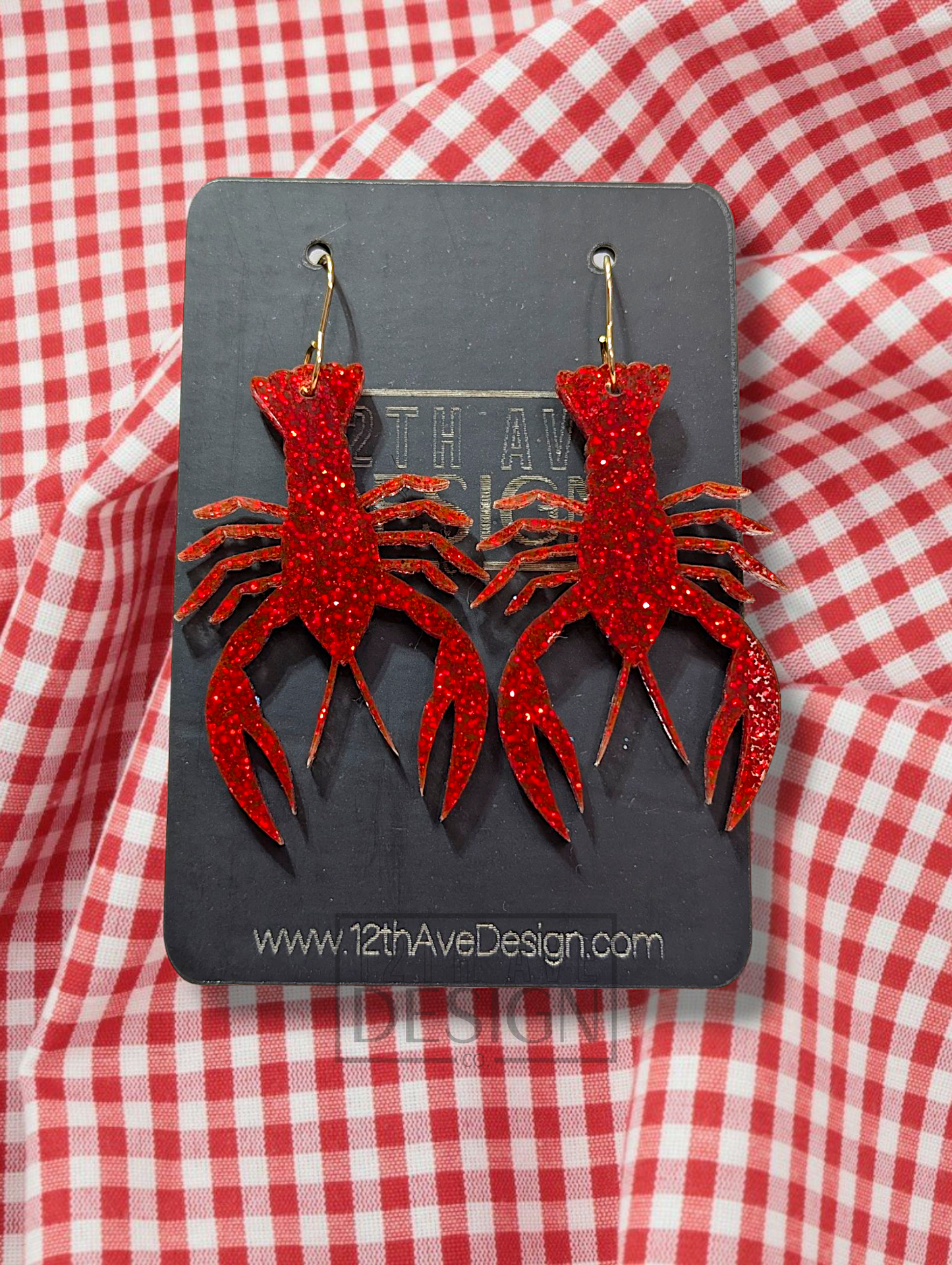 A pair of red glitter-covered acrylic earrings shaped like crawfish, adorned with gold hooks, displayed against a red and white checkered background.