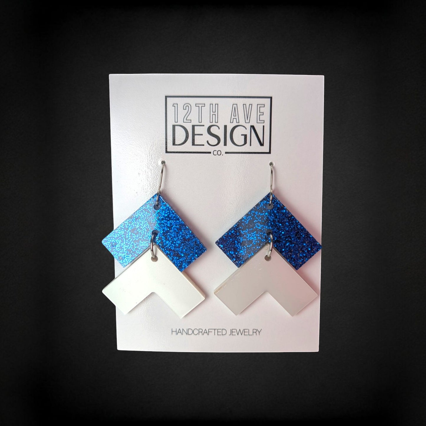 12th Ave Design Co. blue glitter and white chevron drop earrings on surgical steel hooks against a black background.