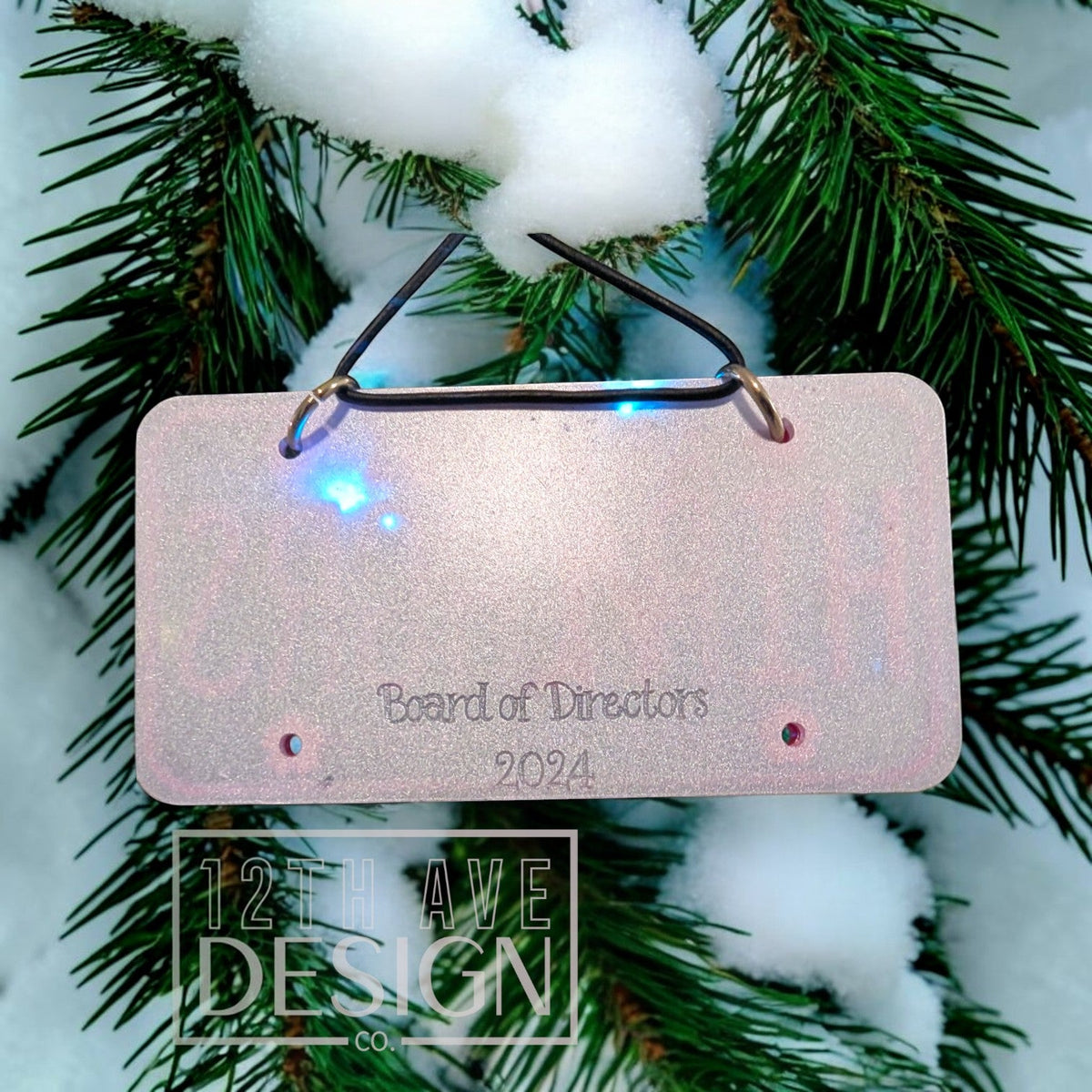 12th Ave Design Co. personalized glittery pink license plate holiday ornament hanging on a snowy pine branch