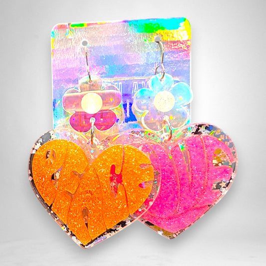 12th Ave Design Co. heart-shaped acrylic earrings with orange and pink glitter lettering and daisy accents.