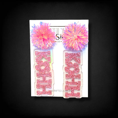 12th Ave Design Co. KOHH varsity bloom earrings featuring pink glitter lettering on clear acrylic with pink pom-poms.