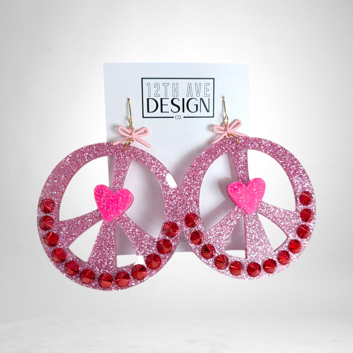 12th Ave Design Co. pink glitter peace sign earrings with red rhinestones, a central heart, and small bow accents.
