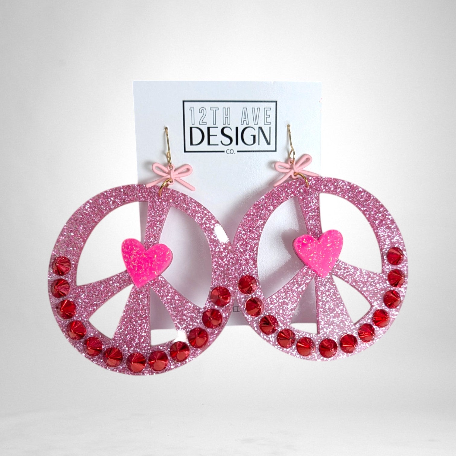 12th Ave Design Co. pink glitter peace sign earrings with red rhinestones, a central heart, and small bow accents.