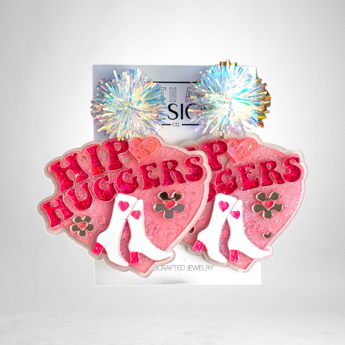 12th Ave Design Co. custom pink heart-shaped resin earrings featuring white cowboy boots and iridescent pom-poms.
