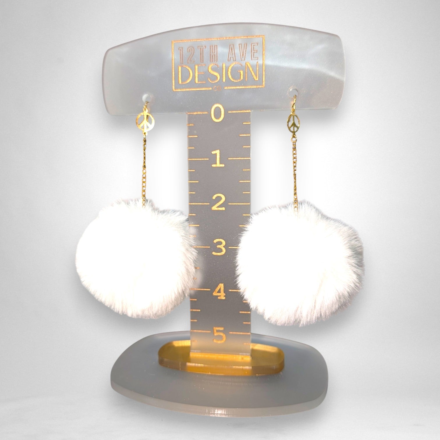 12th Ave Design Co. white fluffy pompom earrings featuring gold chains and peace sign charms on a display stand.