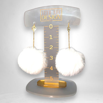 12th Ave Design Co. white fluffy pompom earrings featuring gold chains and peace sign charms on a display stand.