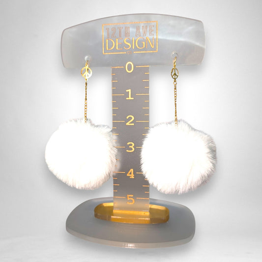 12th Ave Design Co. white fluffy pompom earrings featuring gold chains and peace sign charms on a display stand.