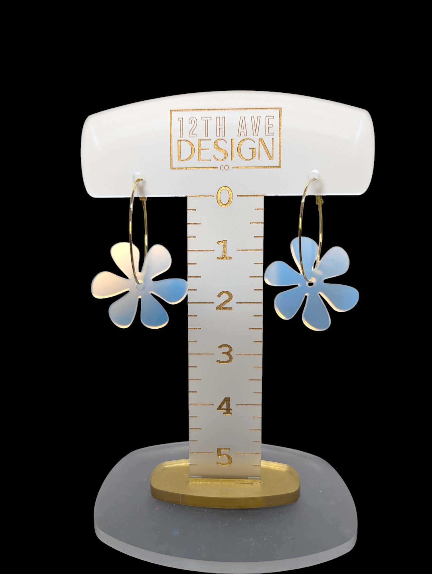 A pair of acrylic flower earrings suspended from a stainless steel hoop, displayed on a measurement ruler marked with '12th ave design'.