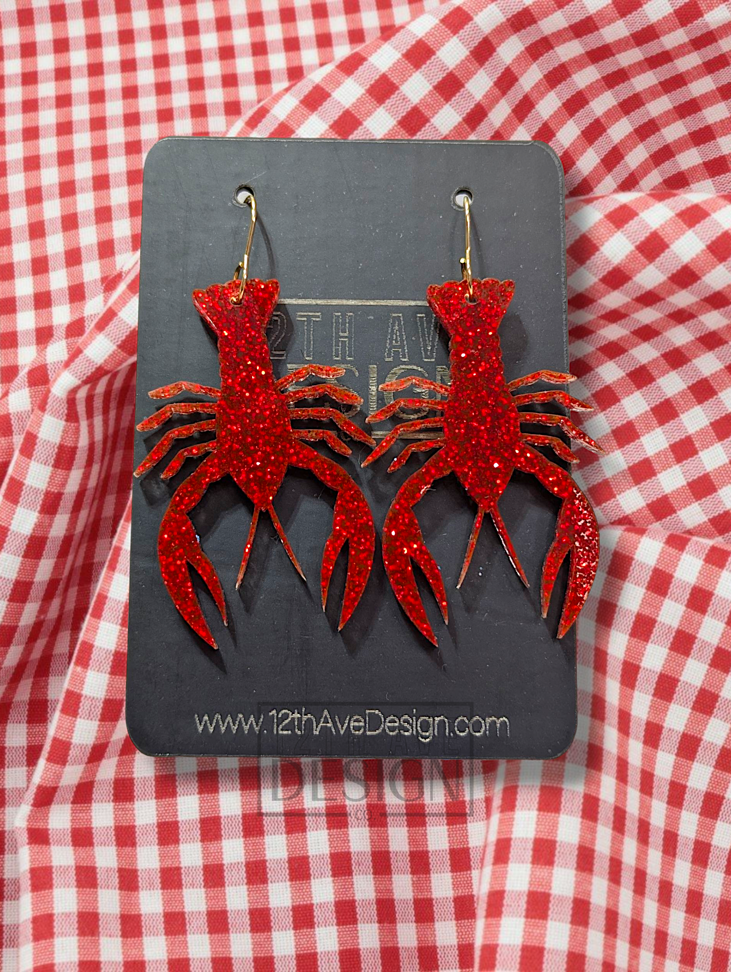 A pair of red glitter-covered acrylic earrings shaped like crawfish, adorned with gold hooks, displayed against a red and white checkered background.