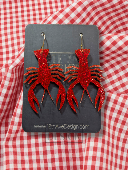A pair of red glitter-covered acrylic earrings shaped like crawfish, adorned with gold hooks, displayed against a red and white checkered background.